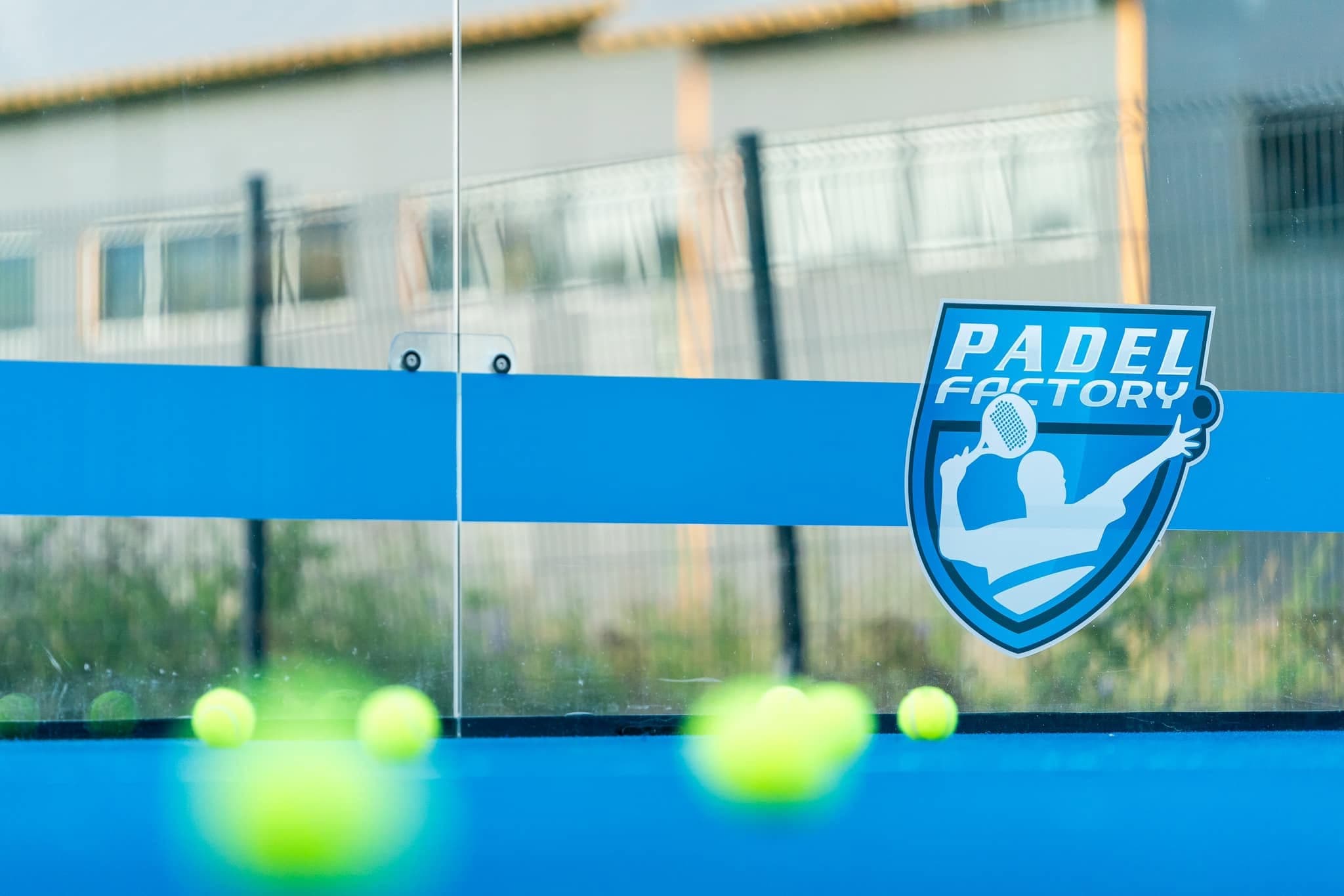 Logo Padel Factory na szklanej bandzie kortu, piłki na niebieskiej nawierzchni